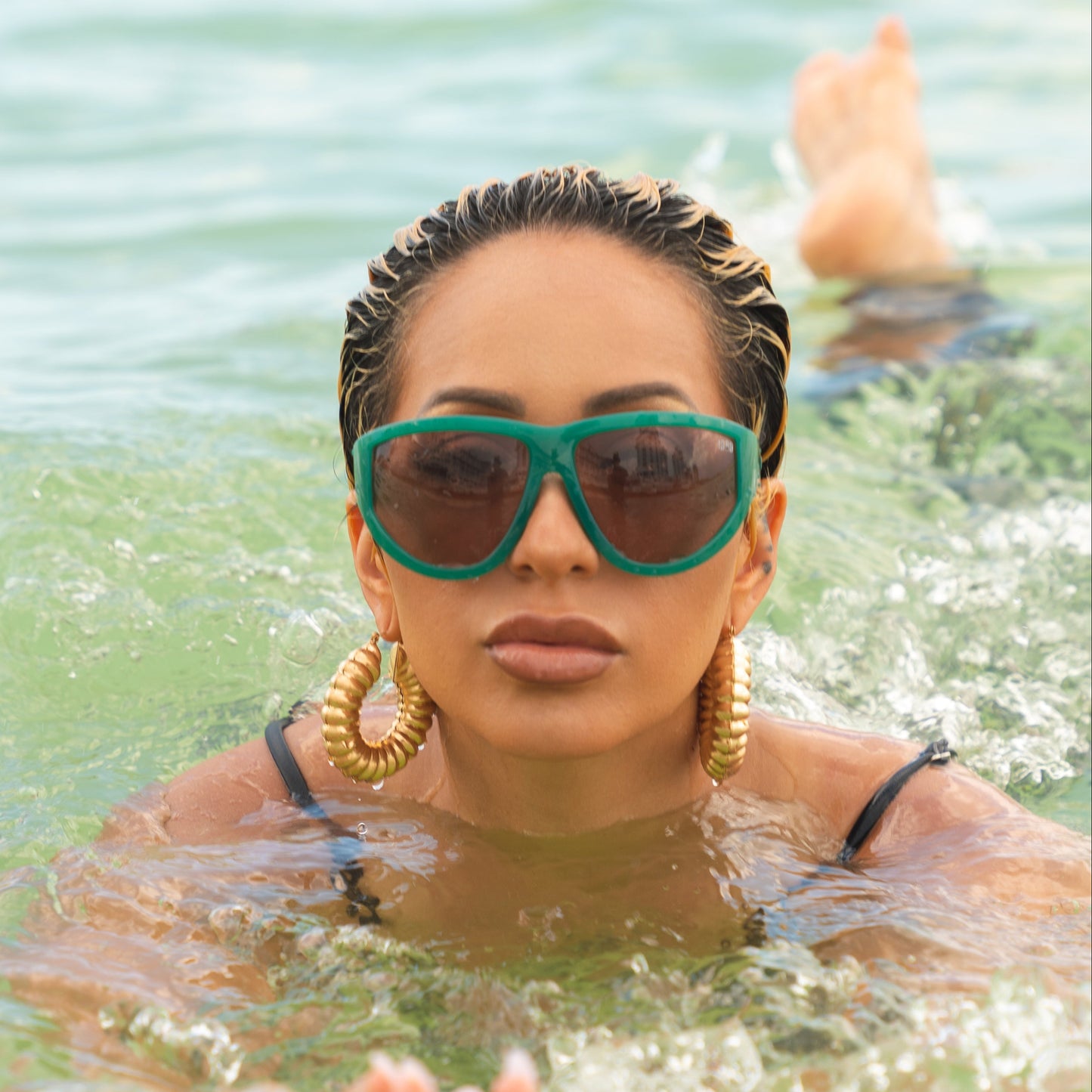 Model is in the water wearing oversized emerald green sunglasses with blush pink lenses and bold curved frame.