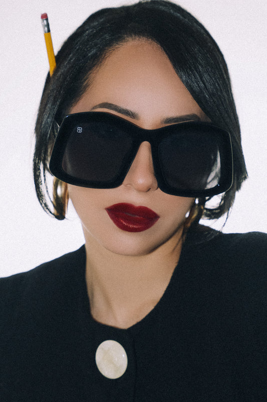 Person wearing large black sunglasses with a pencil on their head against a white background