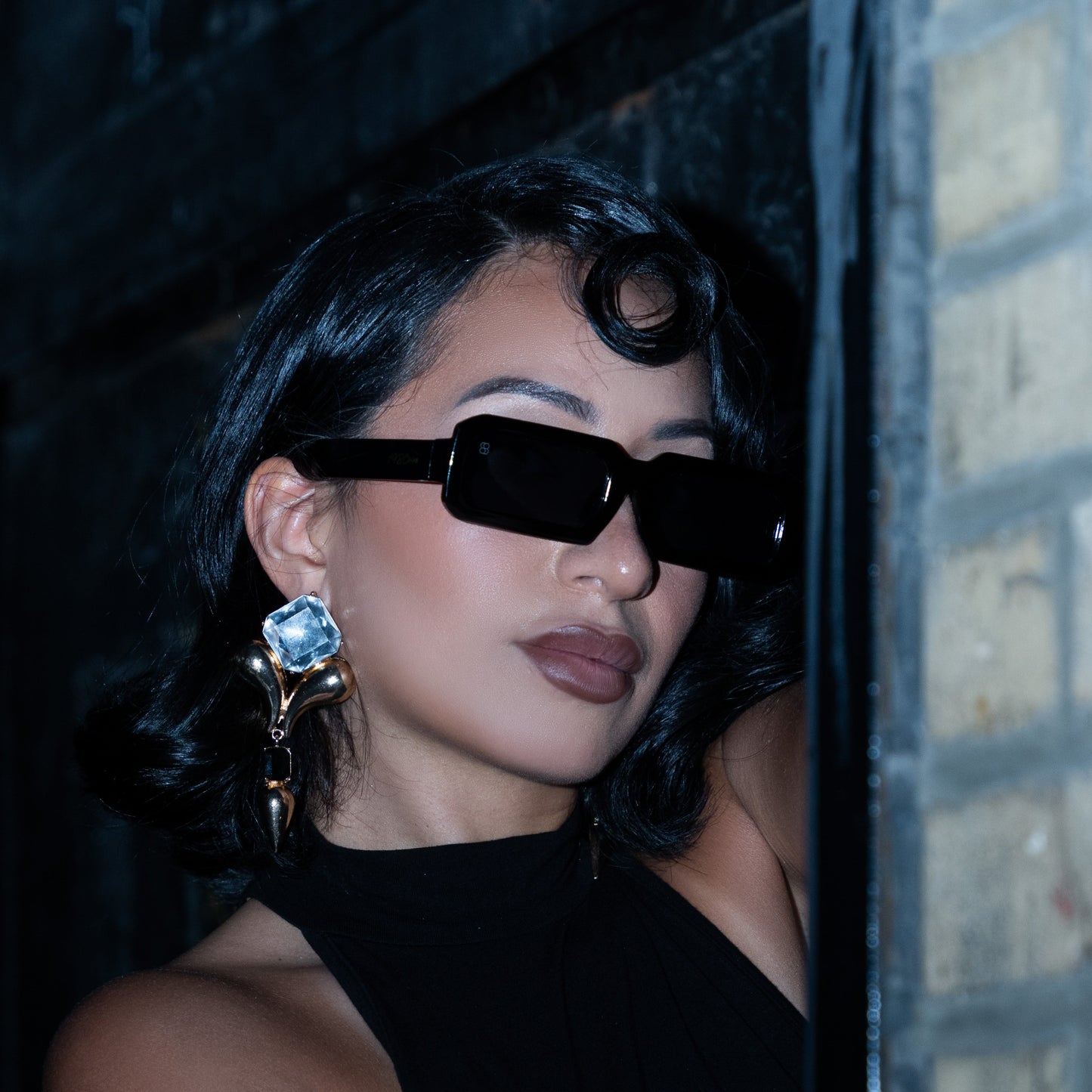 Woman wearing black 1980nine sunglasses and a black dress, standing against a textured wall.