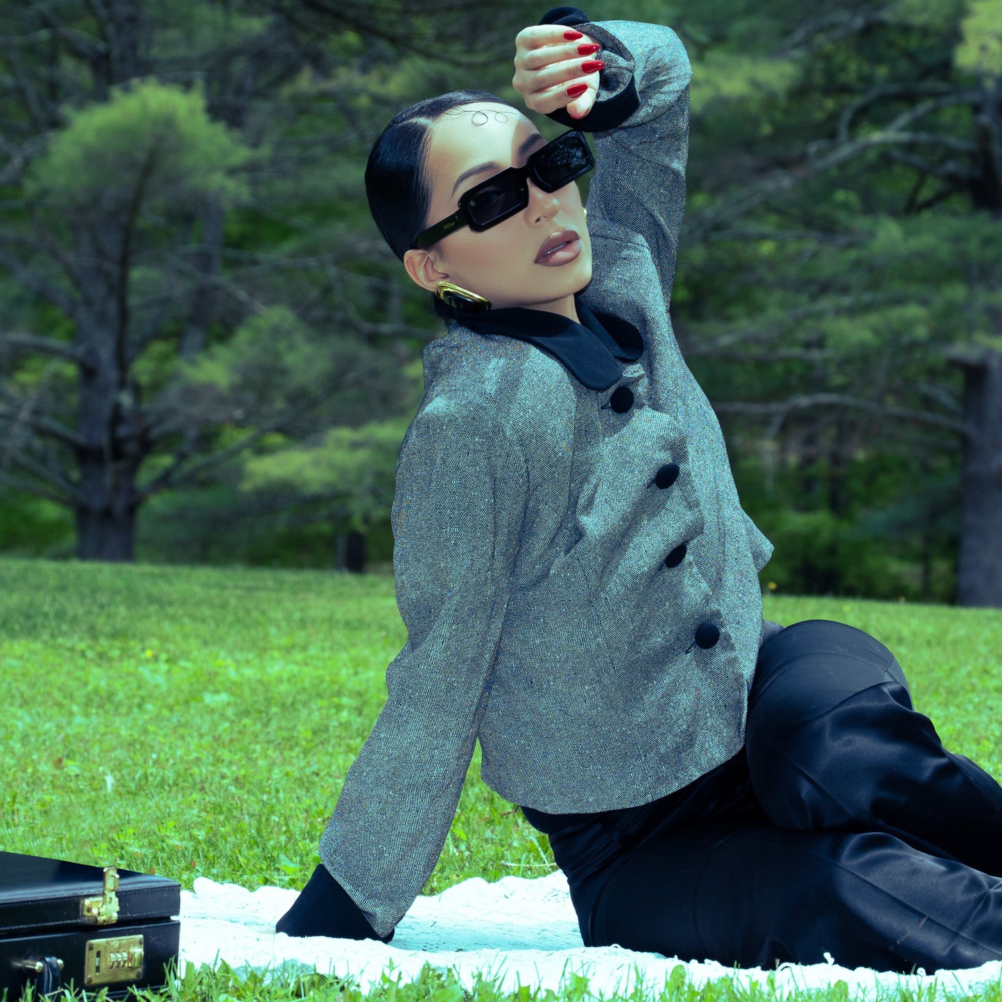 Person in gray coat and sunglasses sitting on grass with trees in the background