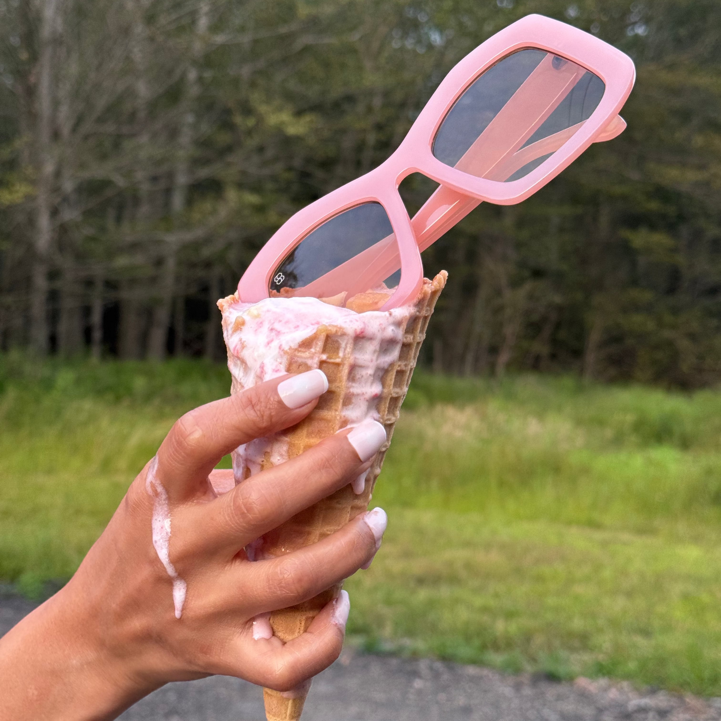 Person holding an ice cream cone with pink sunglasses on top, outdoors.