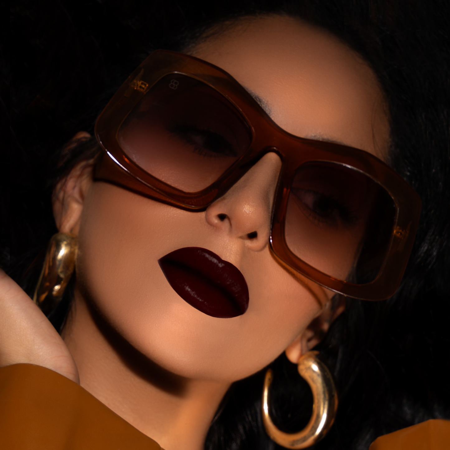 Close-up of a person wearing large 1980nine sunglasses and gold hoop earrings with a dark background