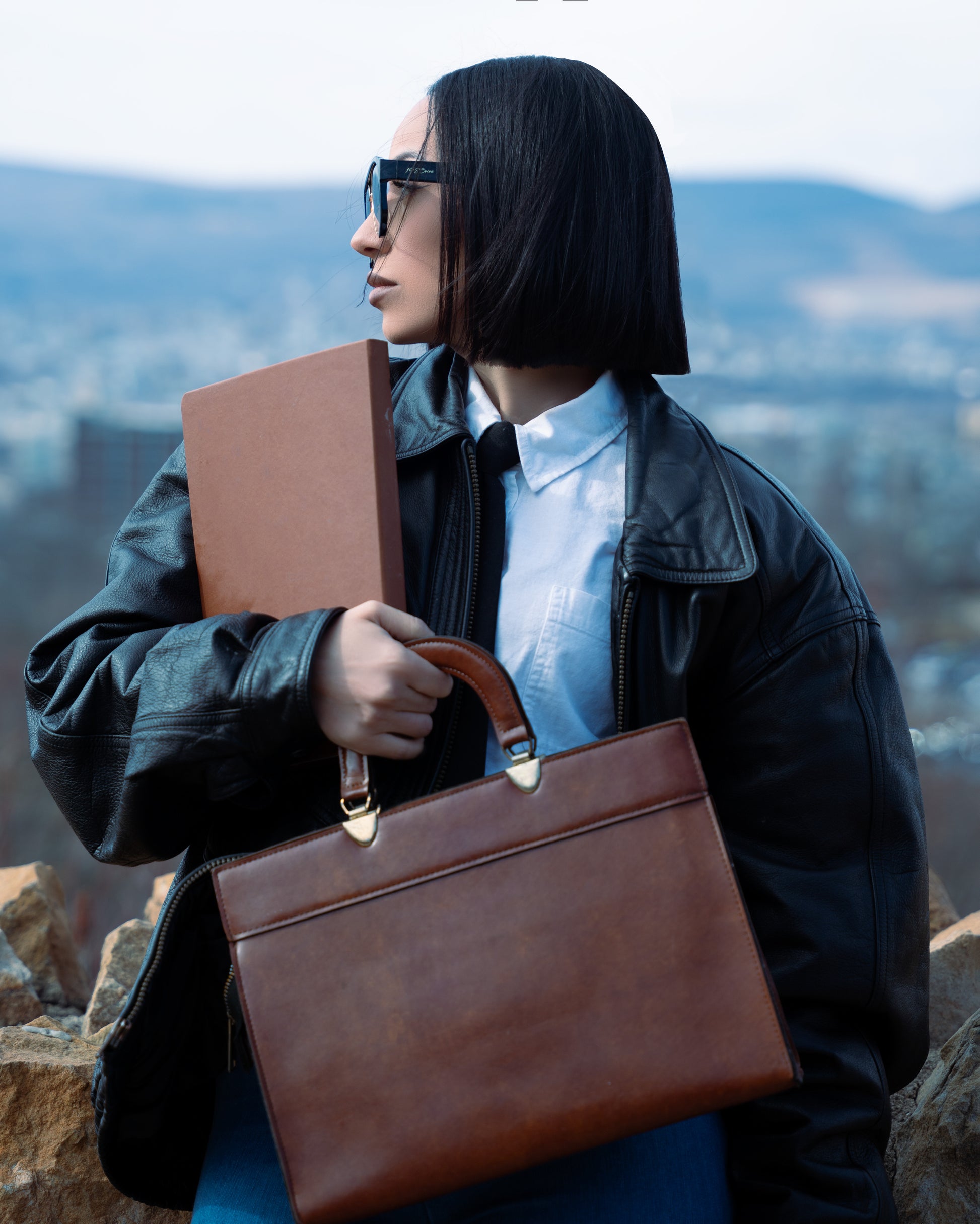 Woman holding a brown leather briefcase with a scenic background wearing 1980nine sunglasses