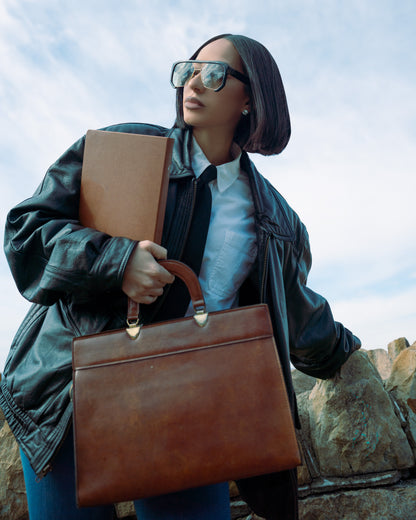 Marissa Roche holding a brown leather briefcase against a natural background wearing 1980nine sunglasses