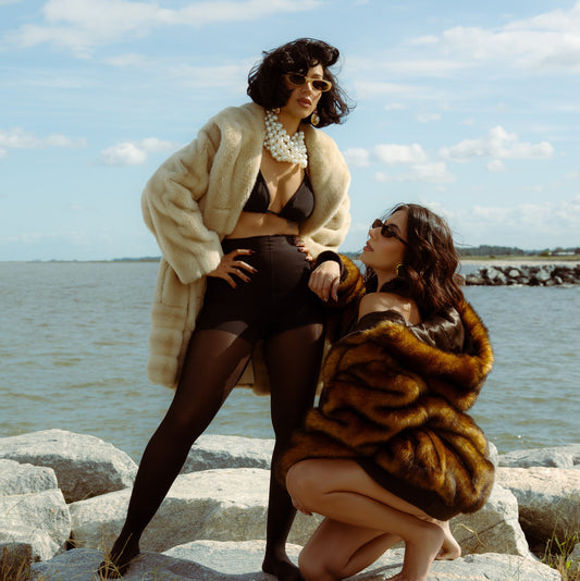 Two women in fur coats by a body of water on a sunny day.