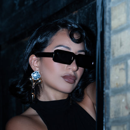 Woman wearing black 1980nine sunglasses and a black dress, standing against a textured wall.