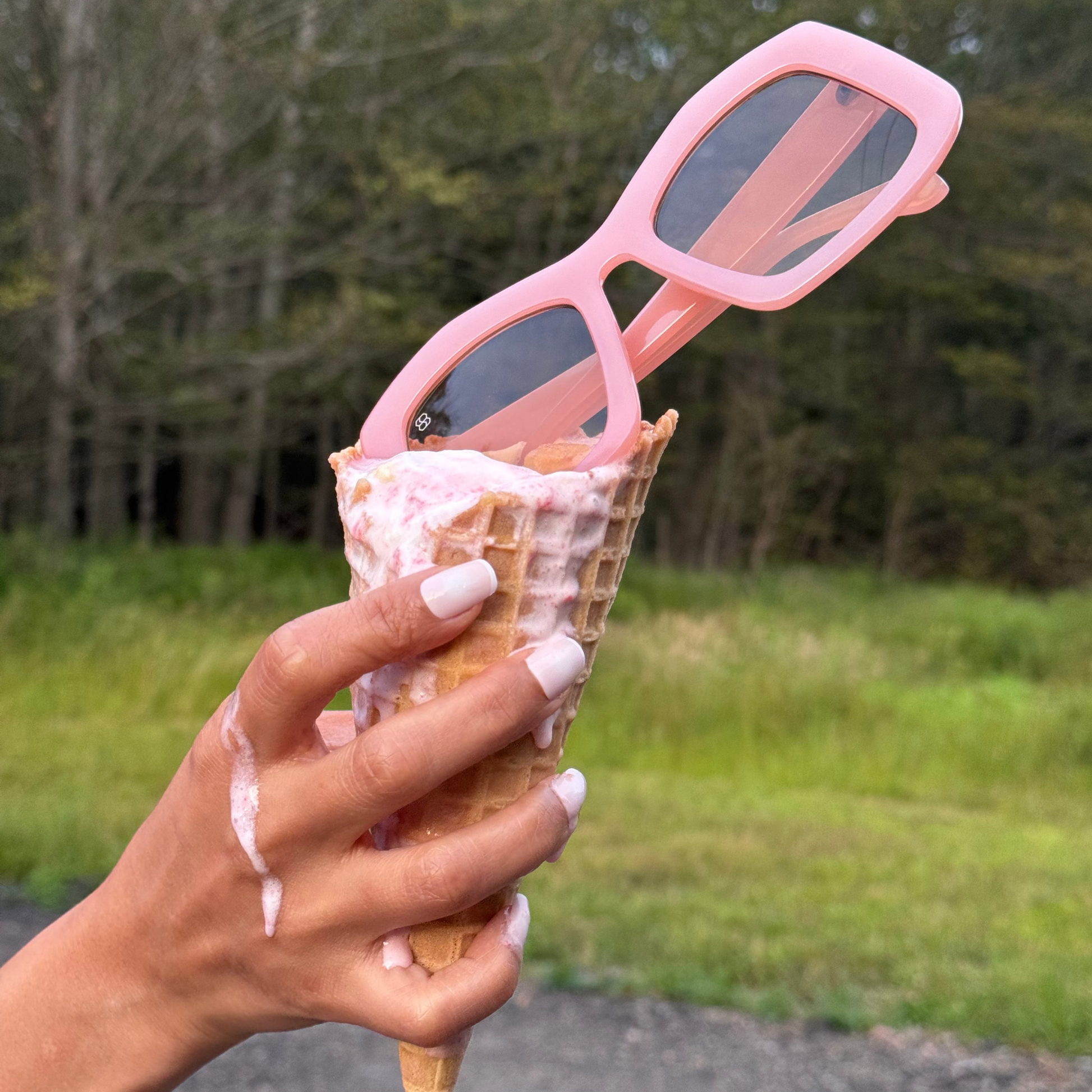 Person holding an ice cream cone with pink sunglasses on top, outdoors.