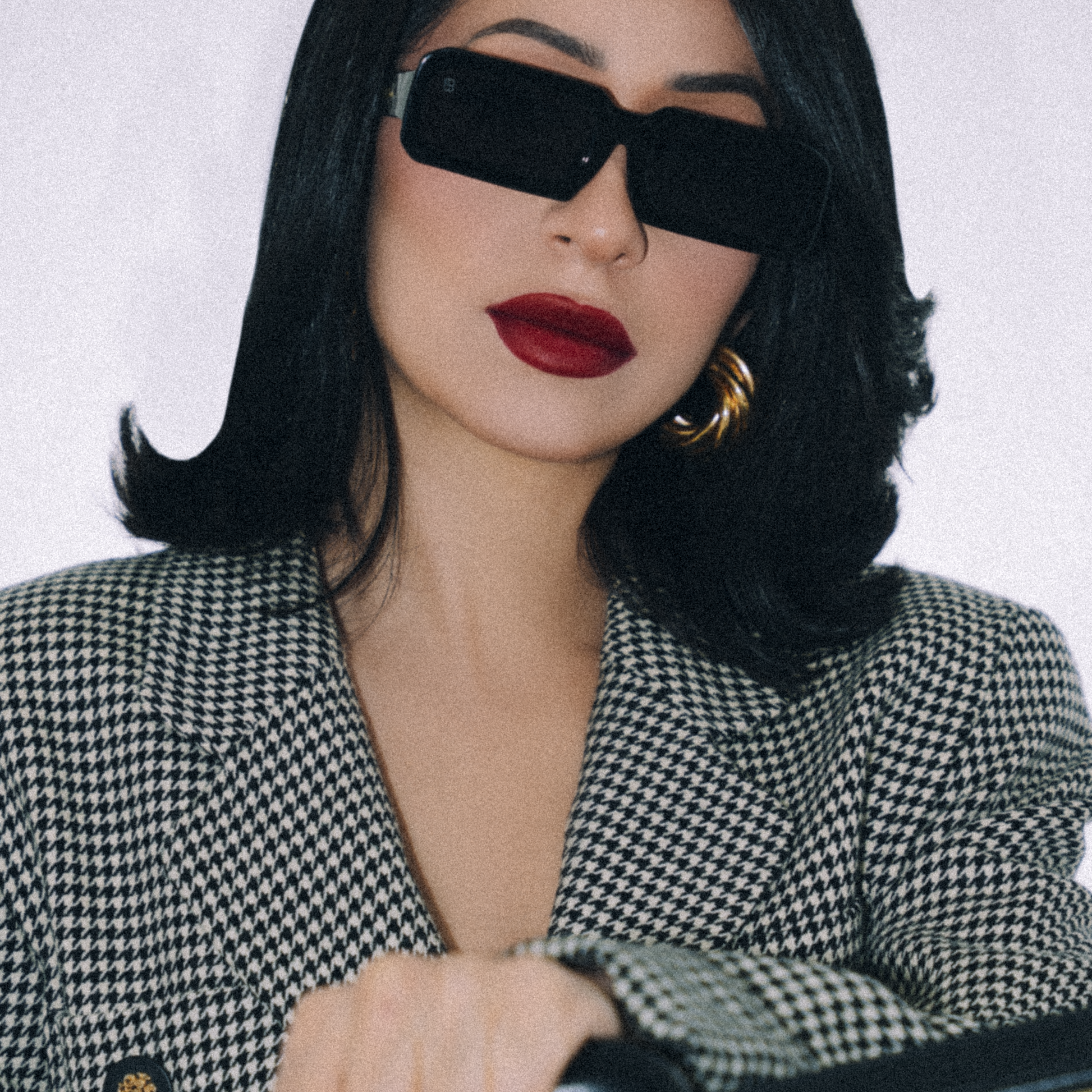 Woman in a checkered blazer and sunglasses holding a suitcase against a white background