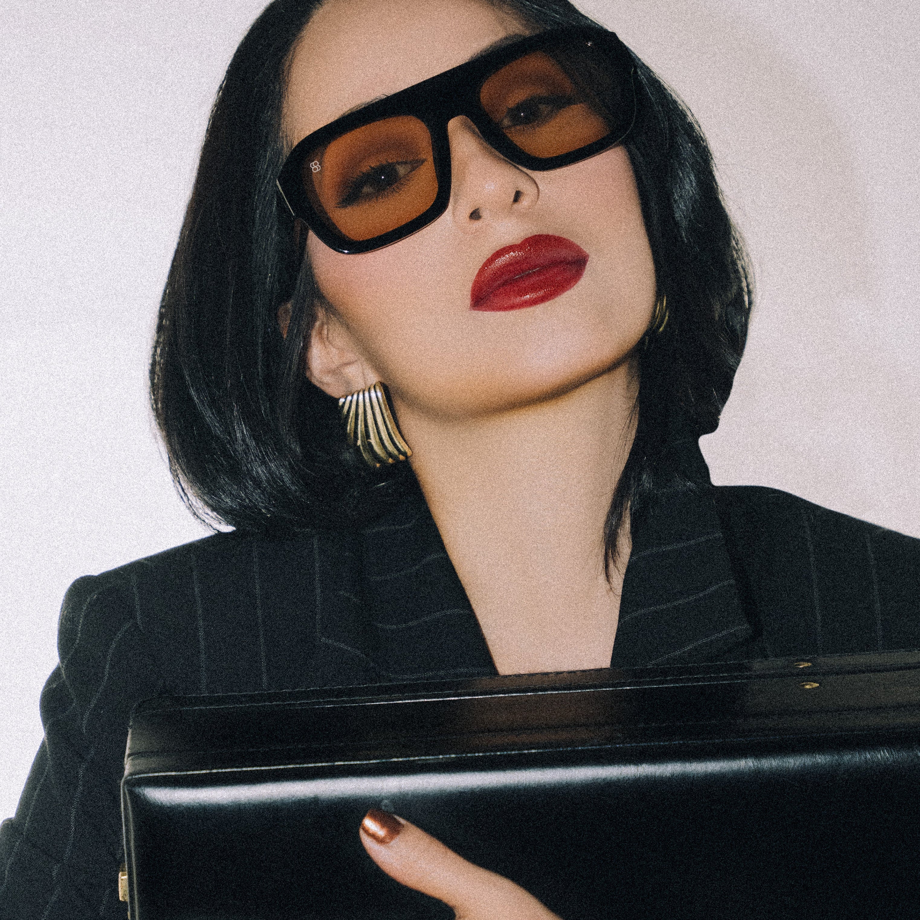 Woman wearing large 1980nine sunglasses and a black outfit, holding a black clutch against a white background