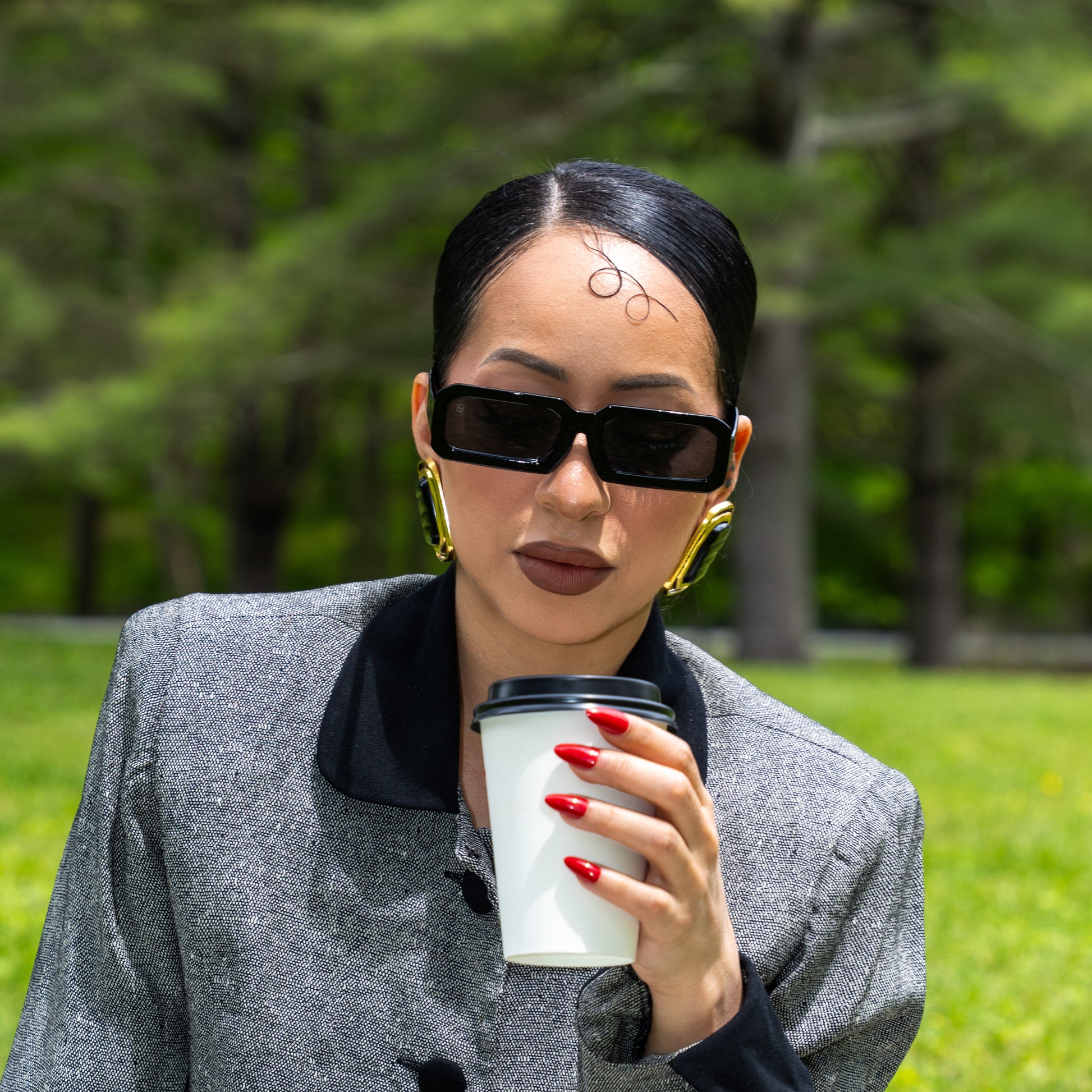 Woman in a gray coat holding a coffee cup outdoors with greenery in the background wearing 1980nine sunglasses